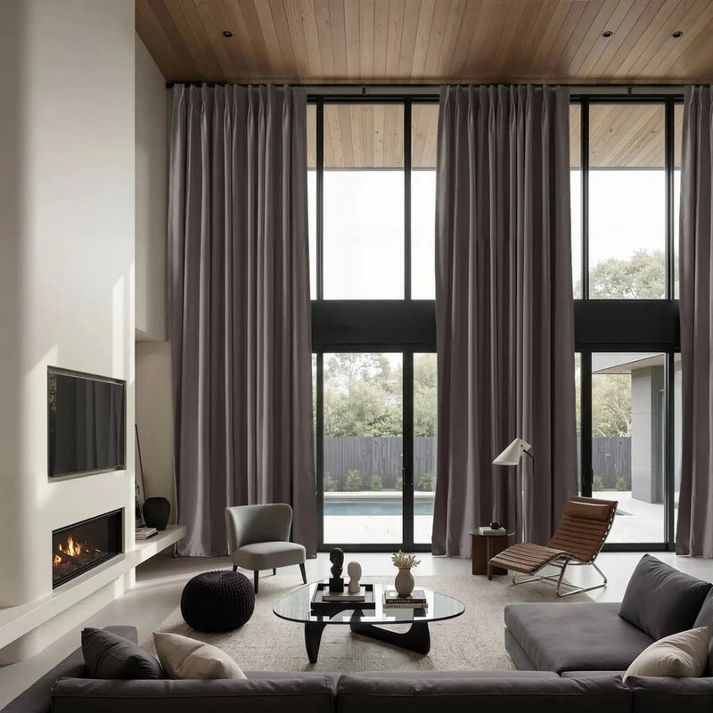 A modern interior design photograph showcasing a spacious living room with expansive grey drapes on floor-to-ceiling windows, a built-in linear fireplace below a wall-mounted TV, and a warm wooden ceiling, leading to an outdoor swimming pool area
