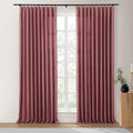 Photograph of two dusty rose pinch-pleat curtains on a black rod in a modern room, framing a bright window revealing green foliage, with a fluted wood side table holding a black vase and brass candle