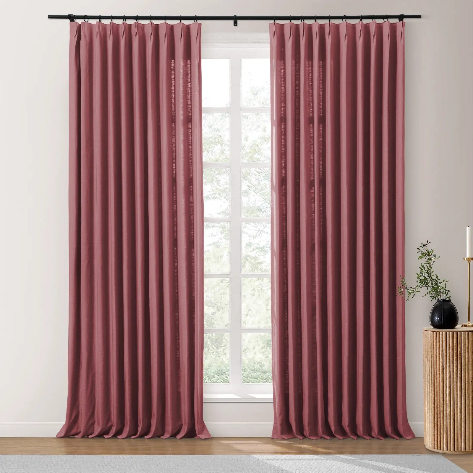 Photograph of two dusty rose pinch-pleat curtains on a black rod in a modern room, framing a bright window revealing green foliage, with a fluted wood side table holding a black vase and brass candle