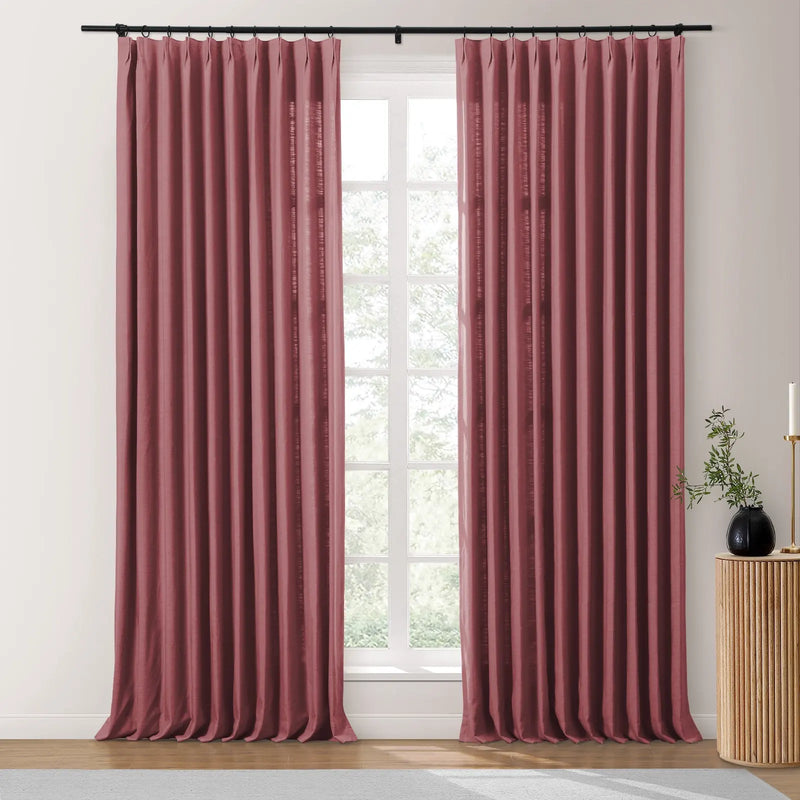 Photograph of two dusty rose pinch-pleat curtains on a black rod in a modern room, framing a bright window revealing green foliage, with a fluted wood side table holding a black vase and brass candle
