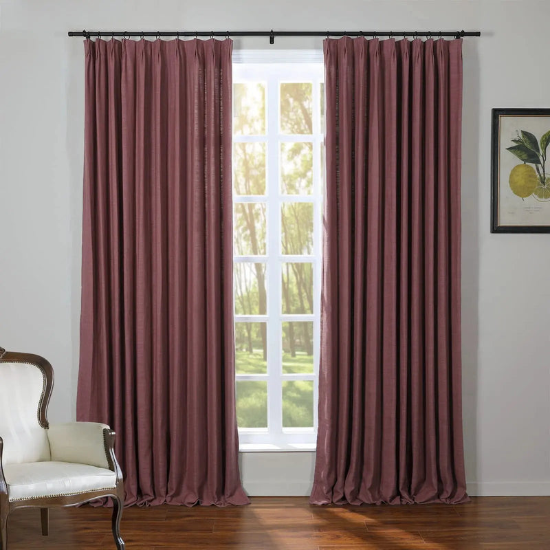 An interior scene featuring dusty rose pinch-pleat curtains on a black rod framing a multi-pane window revealing sunlit green trees, alongside a white antique armchair with decorative nailhead trim and a botanical lemon print on the wall