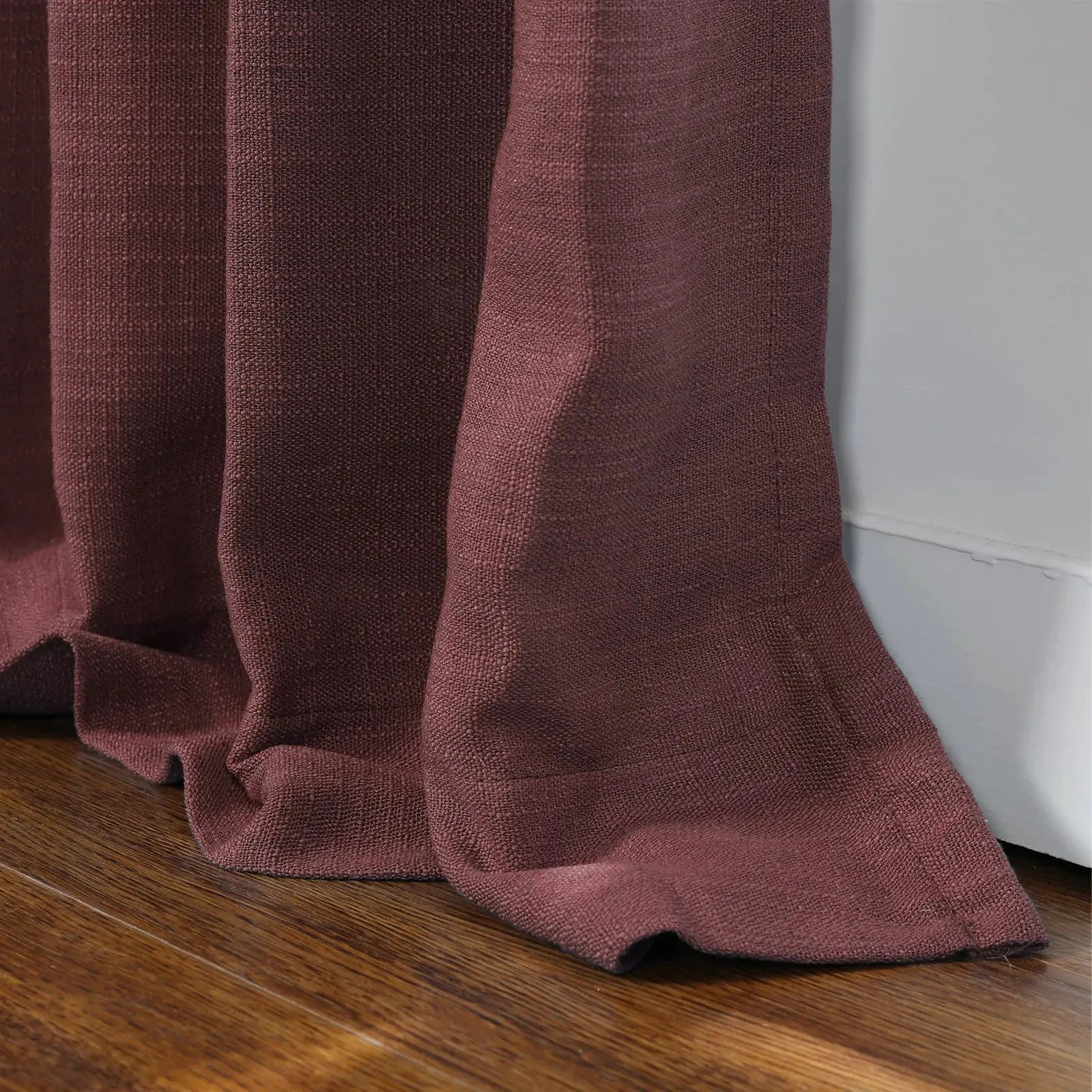 Photograph of a deep burgundy textured curtain with elegant folds pooling on a polished dark wooden floor next to a white baseboard