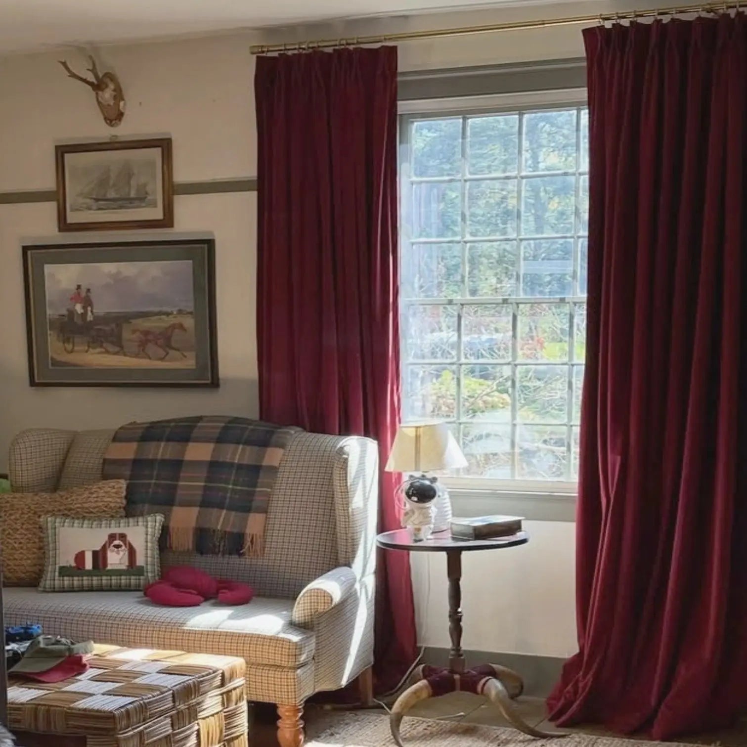 Photograph of a traditional living room with vivid red velvet curtains framing a mullioned window a plaid wingback loveseat adorned with a basset hound pillow and an unusual astronaut figurine lamp on a table with horn legs