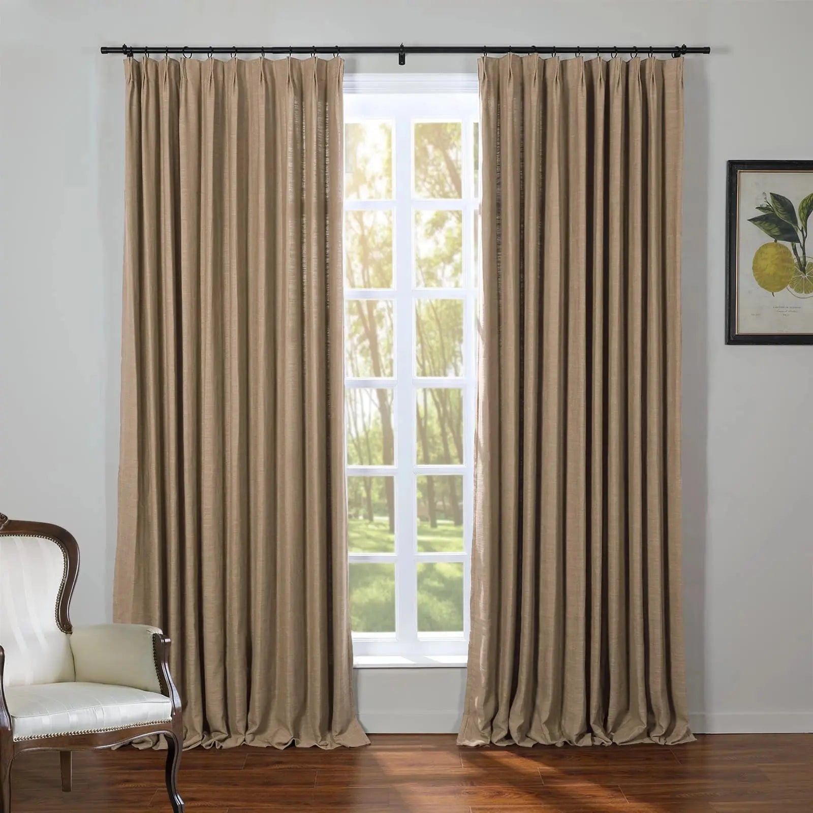 A room interior showcasing medium brown pleated drapes on a black rod framing a white multi-pane window overlooking sunlit green trees, with a classic white upholstered armchair on the left and a framed lemon botanical print on the right wall