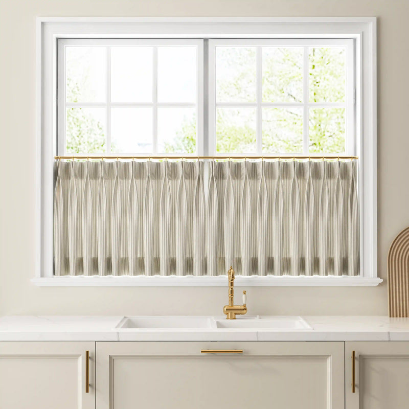 A modern kitchen featuring a window with pleated striped cafe curtains on a brass rod above a white double sink with a sleek gold faucet set into a white quartz countertop