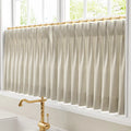 Photograph of a kitchen window featuring pleated beige and white striped cafe curtains on a gold rod above a polished brass vintage faucet with a white ceramic handle offering a leafy green outdoor view