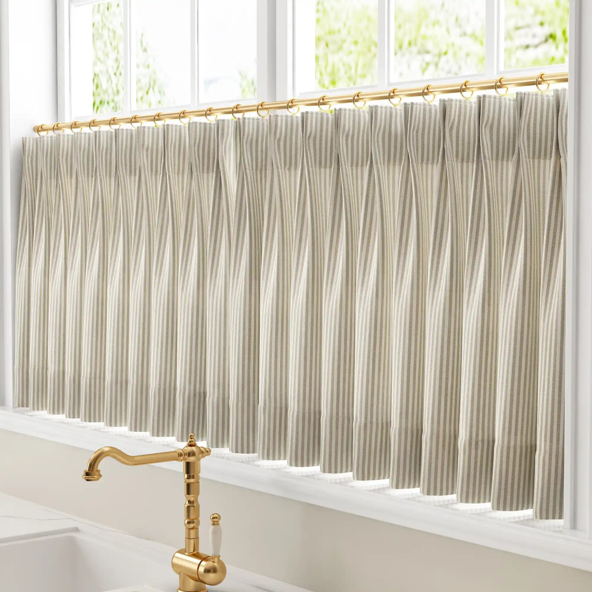 Photograph of a kitchen window featuring pleated beige and white striped cafe curtains on a gold rod above a polished brass vintage faucet with a white ceramic handle offering a leafy green outdoor view