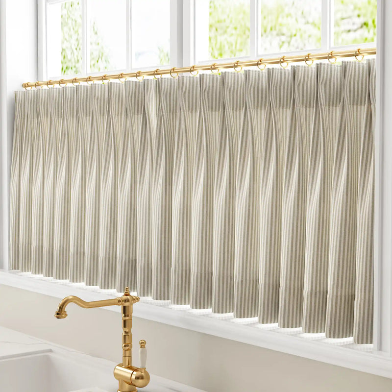 Photograph of a kitchen window featuring pleated beige and white striped cafe curtains on a gold rod above a polished brass vintage faucet with a white ceramic handle offering a leafy green outdoor view