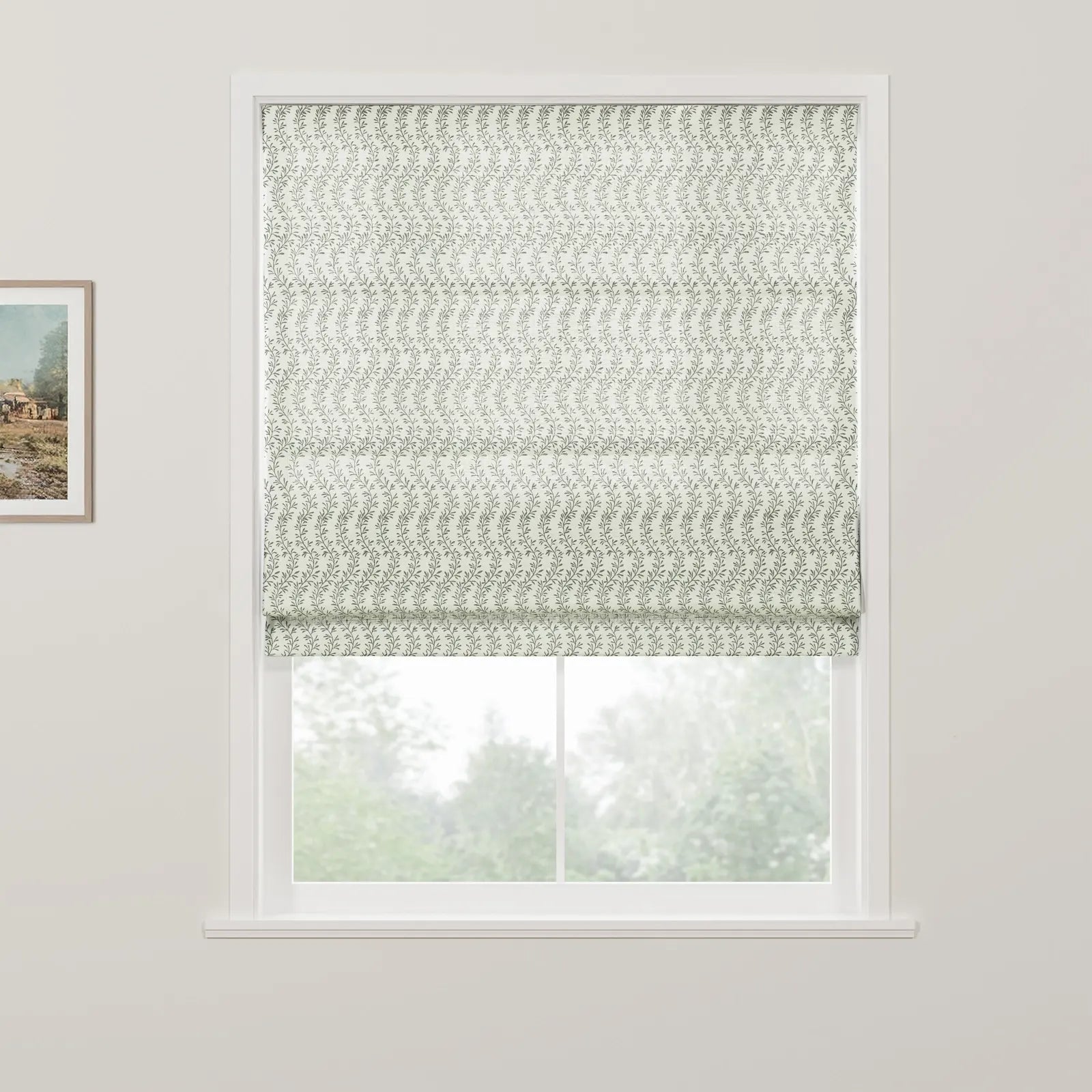 A white Roman blind with a delicate green leaf pattern is partially drawn over a white window frame, offering a blurred view of lush green trees beyond, with a rustic framed landscape painting visible on the wall