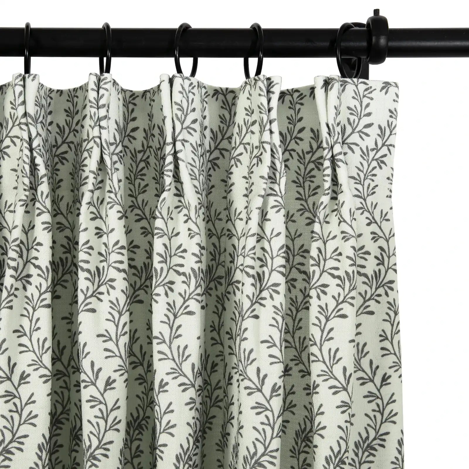 Photograph of a light green pleated curtain featuring a dark gray leafy branch pattern, suspended by black rings from a black curtain rod