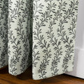 Fabric curtains featuring a light cream woven texture with a repeating dark grey vine pattern, draped in soft vertical pleats and resting on a warm brown wooden floor next to a white baseboard