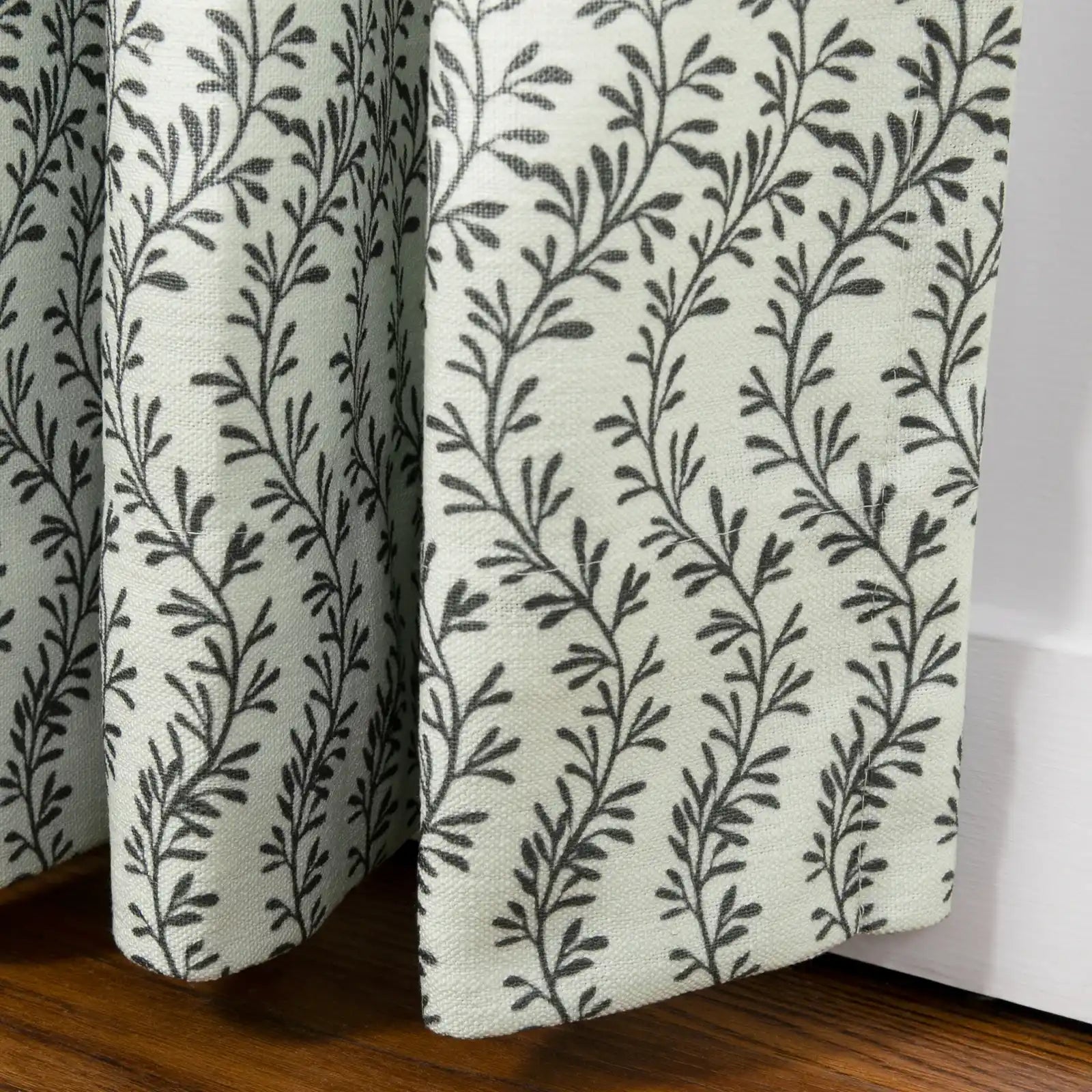 Fabric curtains featuring a light cream woven texture with a repeating dark grey vine pattern, draped in soft vertical pleats and resting on a warm brown wooden floor next to a white baseboard