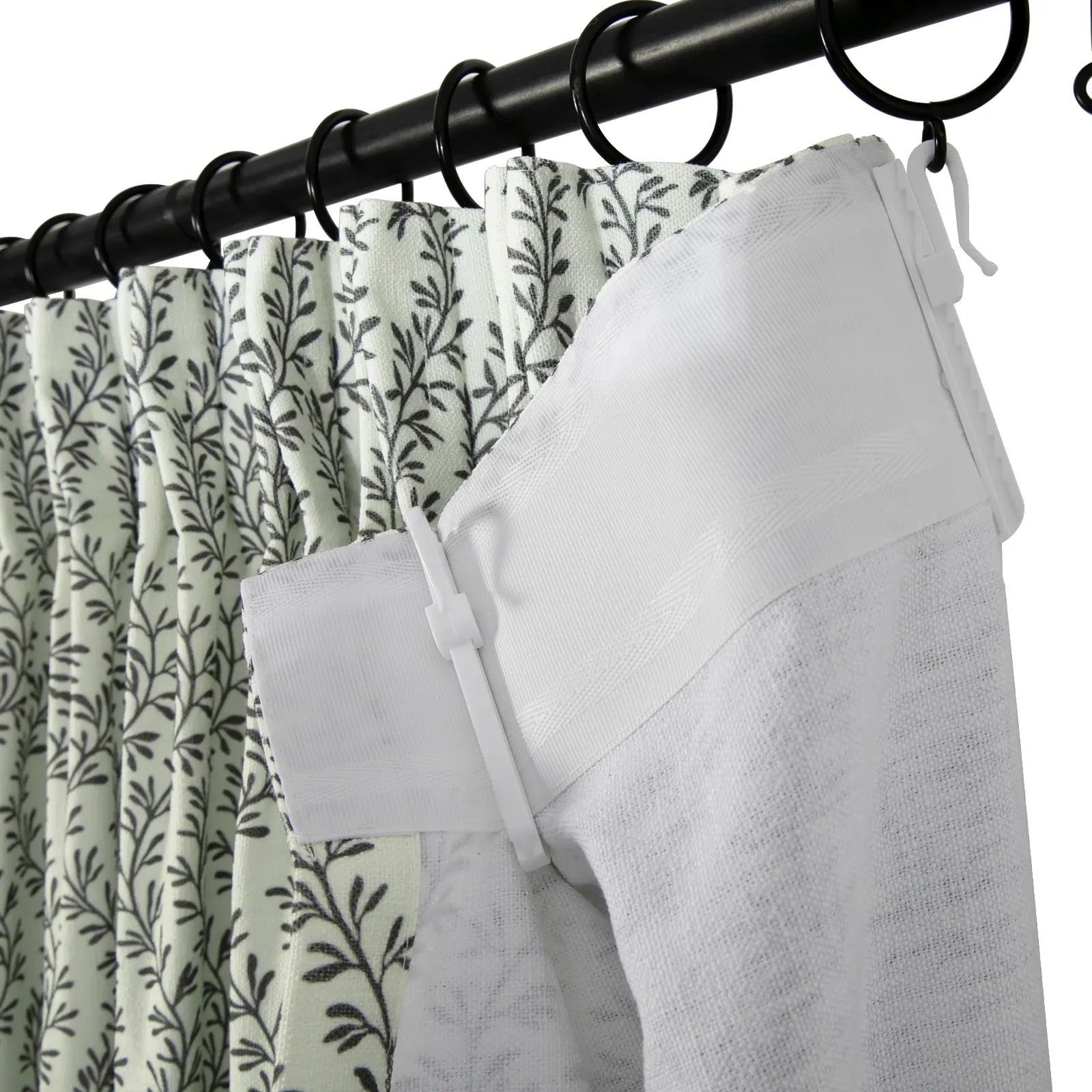 Black curtain rod with rings holding a pleated white curtain featuring a dark green botanical leaf pattern and a solid white textured curtain panel with a herringbone weave joined by a white plastic clip
