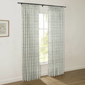 Interior photo showcasing white curtains with a green leaf pattern on a black rod, a window revealing lush green outdoor trees, and a wooden-framed botanical print on a light wall