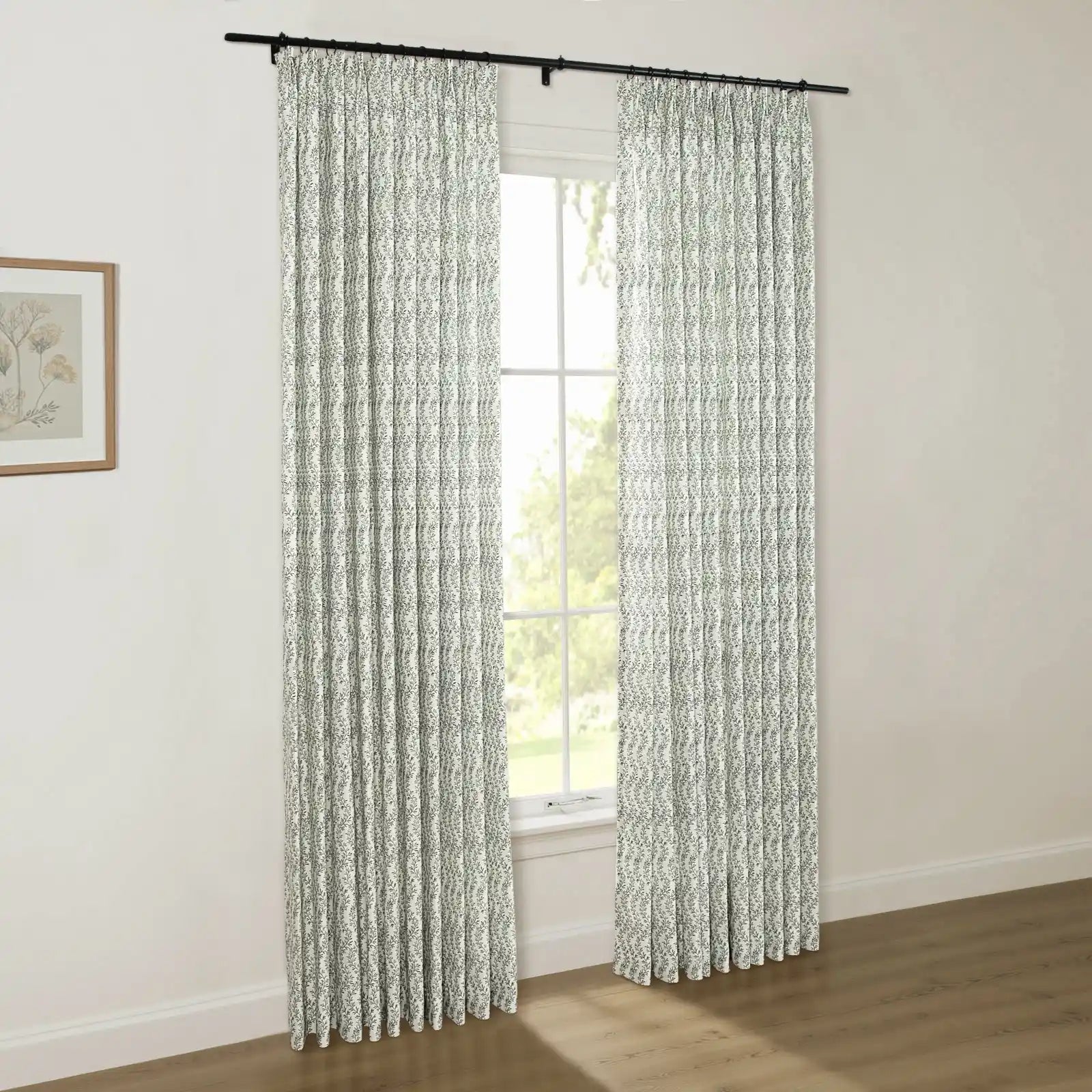 Interior photo showcasing white curtains with a green leaf pattern on a black rod, a window revealing lush green outdoor trees, and a wooden-framed botanical print on a light wall
