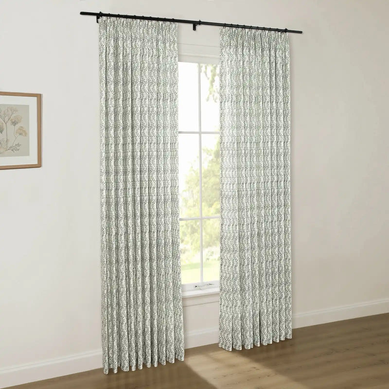 Interior photo showcasing white curtains with a green leaf pattern on a black rod, a window revealing lush green outdoor trees, and a wooden-framed botanical print on a light wall