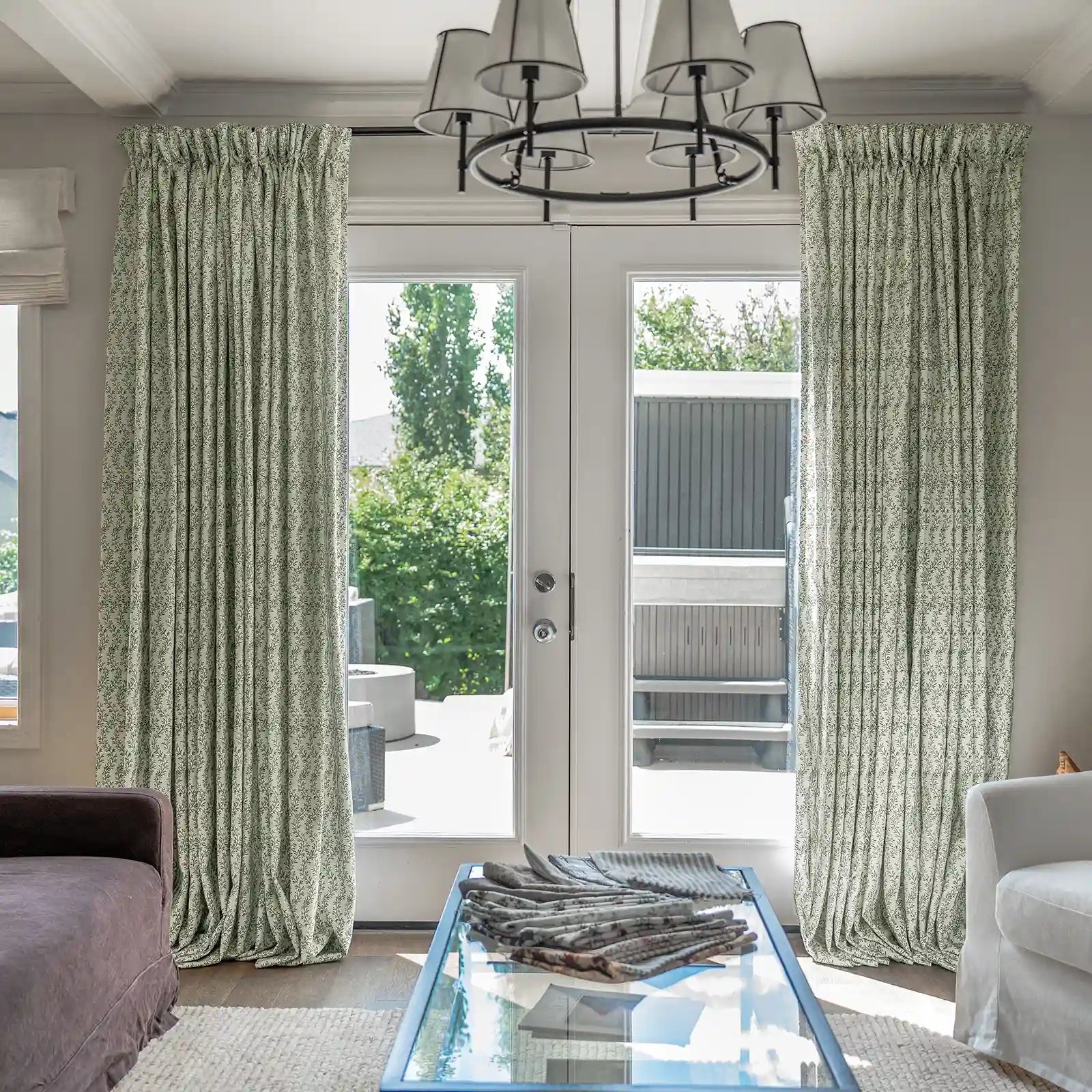 Interior view of a bright living room featuring light green patterned drapes framing white French doors that open to an outdoor patio with a visible hot tub and fire pit, complemented by a black modern chandelier and a glass coffee table displaying fabric samples