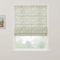 An off-white Roman blind with a subtle grey-green floral vine pattern is partially raised on a white window, revealing blurred green trees outside and a framed rustic landscape painting on the adjacent white wall