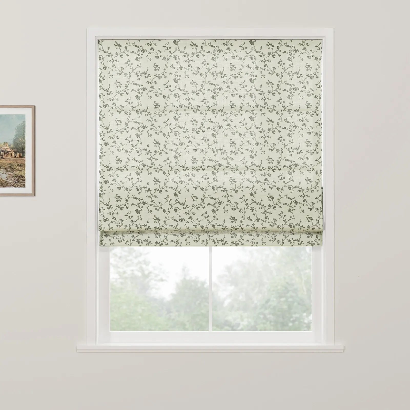 An off-white Roman blind with a subtle grey-green floral vine pattern is partially raised on a white window, revealing blurred green trees outside and a framed rustic landscape painting on the adjacent white wall