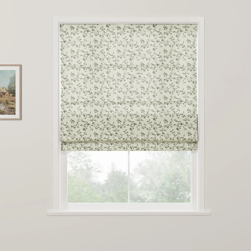 An off-white Roman blind with a subtle grey-green floral vine pattern is partially raised on a white window, revealing blurred green trees outside and a framed rustic landscape painting on the adjacent white wall