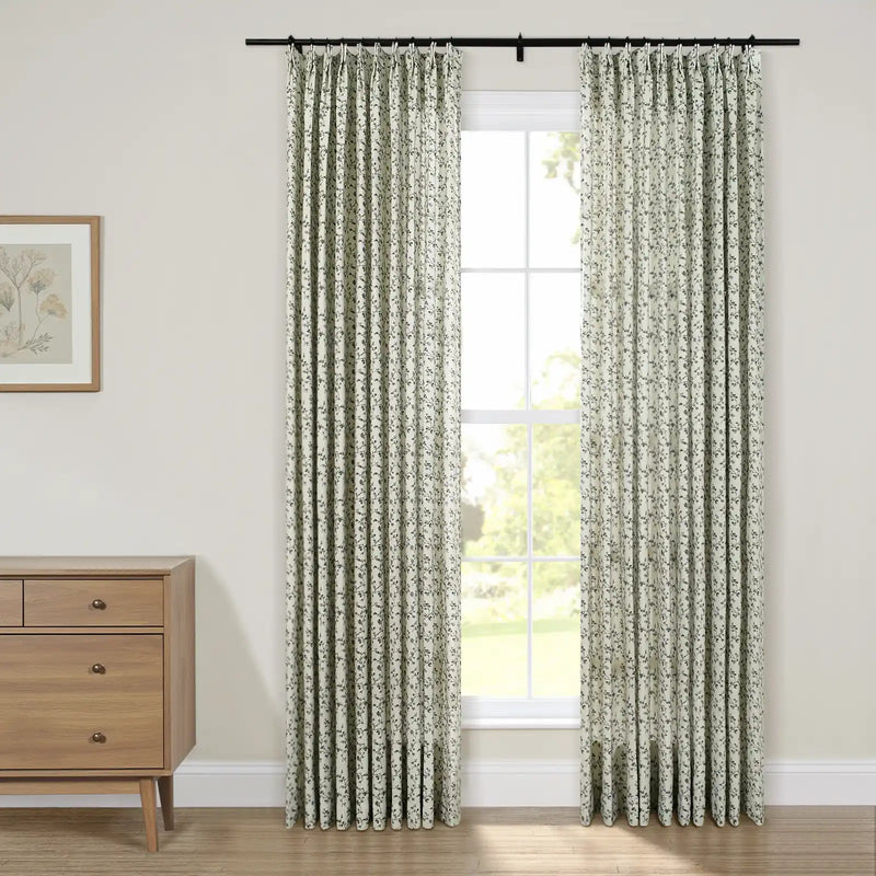 Light sage pleated curtains with a dark floral pattern on a black rod frame a four-pane window overlooking a green outdoor scene, alongside a wooden dresser and framed botanical art