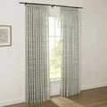 A pair of light floral patterned pinch-pleated curtains on a black rod cover a window, revealing green trees outside, with a framed botanical print on the adjacent white wall