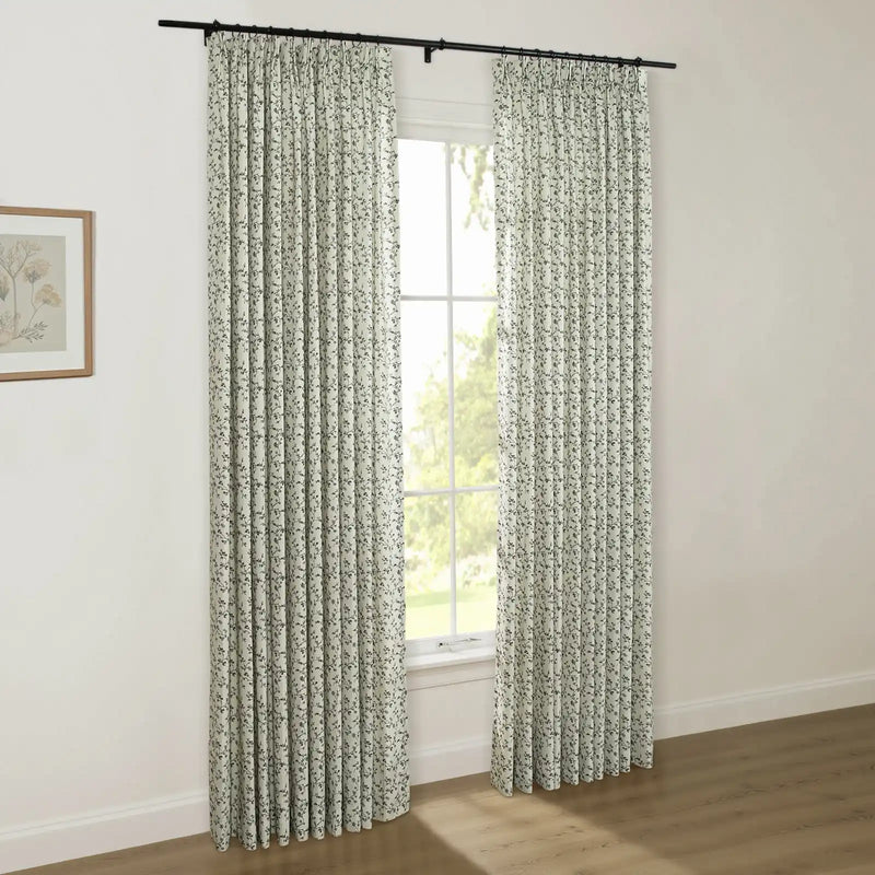 A pair of light floral patterned pinch-pleated curtains on a black rod cover a window, revealing green trees outside, with a framed botanical print on the adjacent white wall