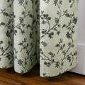 A light sage green curtain featuring a dark green vine and floral pattern displays its woven texture and gathered folds above a warm wooden floor and white baseboard