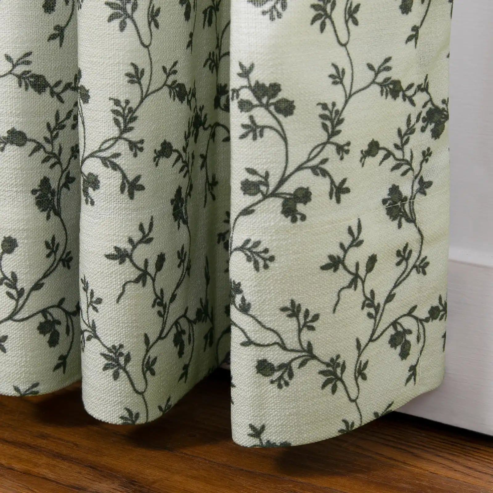 A light sage green curtain featuring a dark green vine and floral pattern displays its woven texture and gathered folds above a warm wooden floor and white baseboard