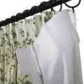Light green fabric curtains featuring a dark green vine pattern hanging on a black rod with rings, revealing a white back lining with plastic clip hooks