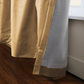 Close-up of a two-tone golden champagne and light gray lined drapery with a subtle textured weave, extending to a dark brown hardwood floor next to a white window sill