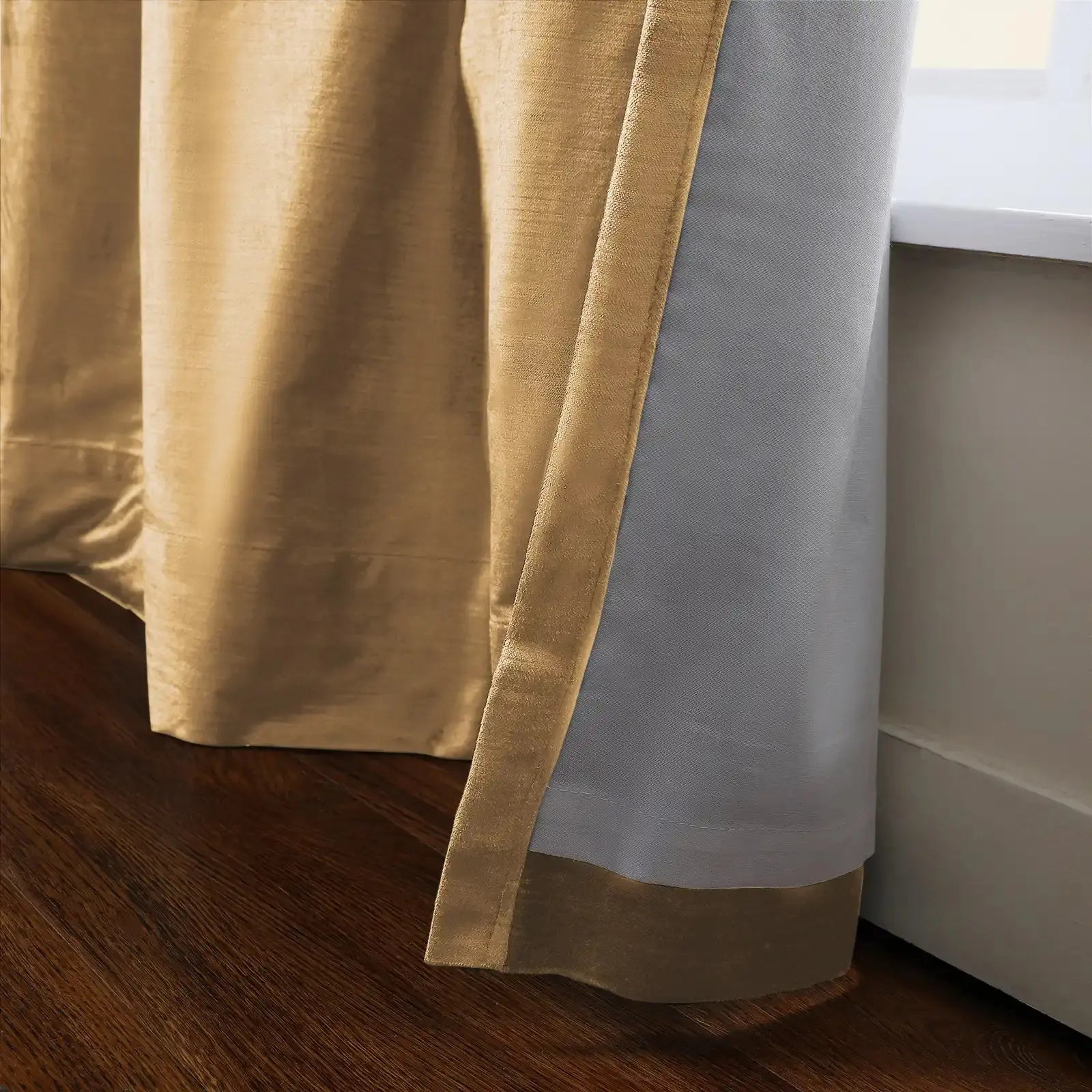 Close-up of a two-tone golden champagne and light gray lined drapery with a subtle textured weave, extending to a dark brown hardwood floor next to a white window sill