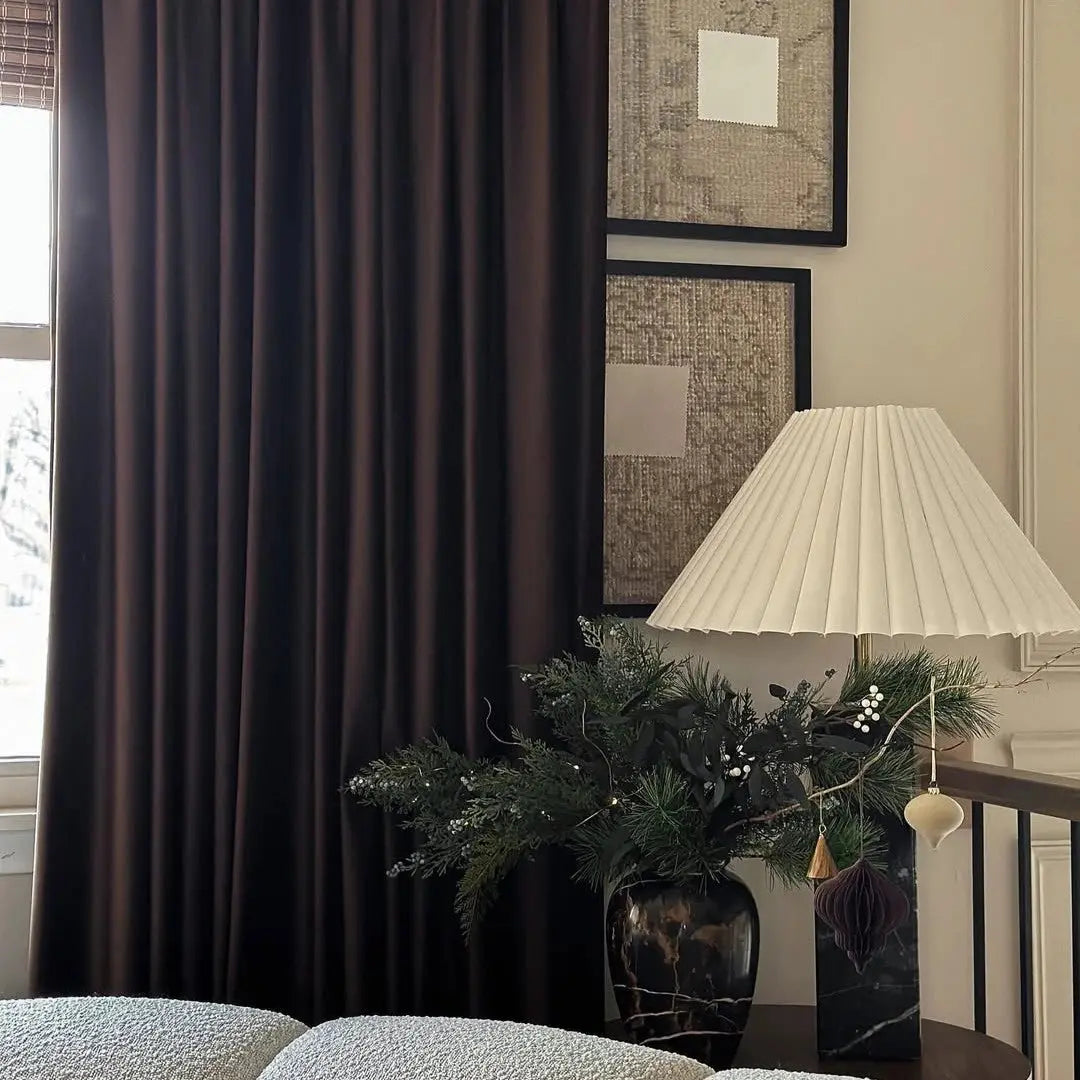 An elegant home interior featuring rich dark brown velvet curtains, a white pleated lamp with a black marble base, and a festive evergreen arrangement adorned with white berries and gold and purple ornaments