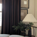An elegant home interior featuring rich dark brown velvet curtains, a white pleated lamp with a black marble base, and a festive evergreen arrangement adorned with white berries and gold and purple ornaments