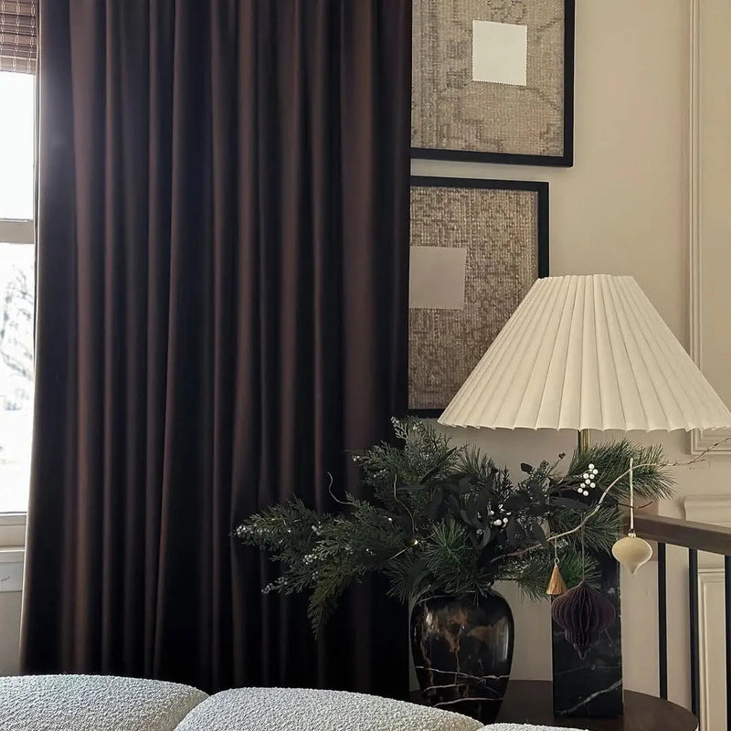 An elegant home interior featuring rich dark brown velvet curtains, a white pleated lamp with a black marble base, and a festive evergreen arrangement adorned with white berries and gold and purple ornaments