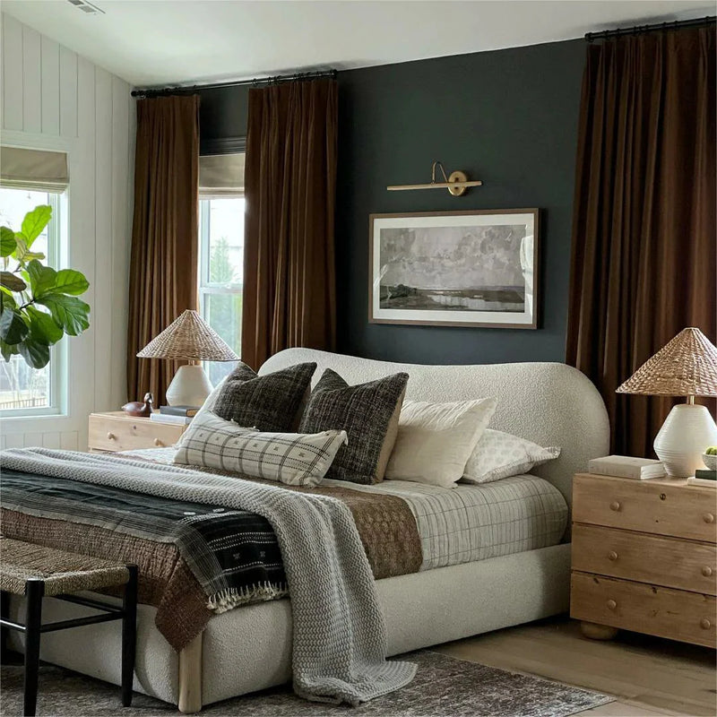 A modern bedroom interior with a dark green accent wall, a curved cream boucle bed layered with neutral plaid and knit textiles, rich brown velvet curtains, and wooden nightstands topped with woven straw lamps