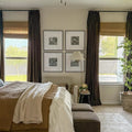 Modern bedroom with a brown quilted bed, layered white and beige throws, a gallery wall of four black and white family photos, large windows with dark brown velvet curtains and bamboo shades overlooking a green backyard, a pleated floor lamp, and a tall potted plant