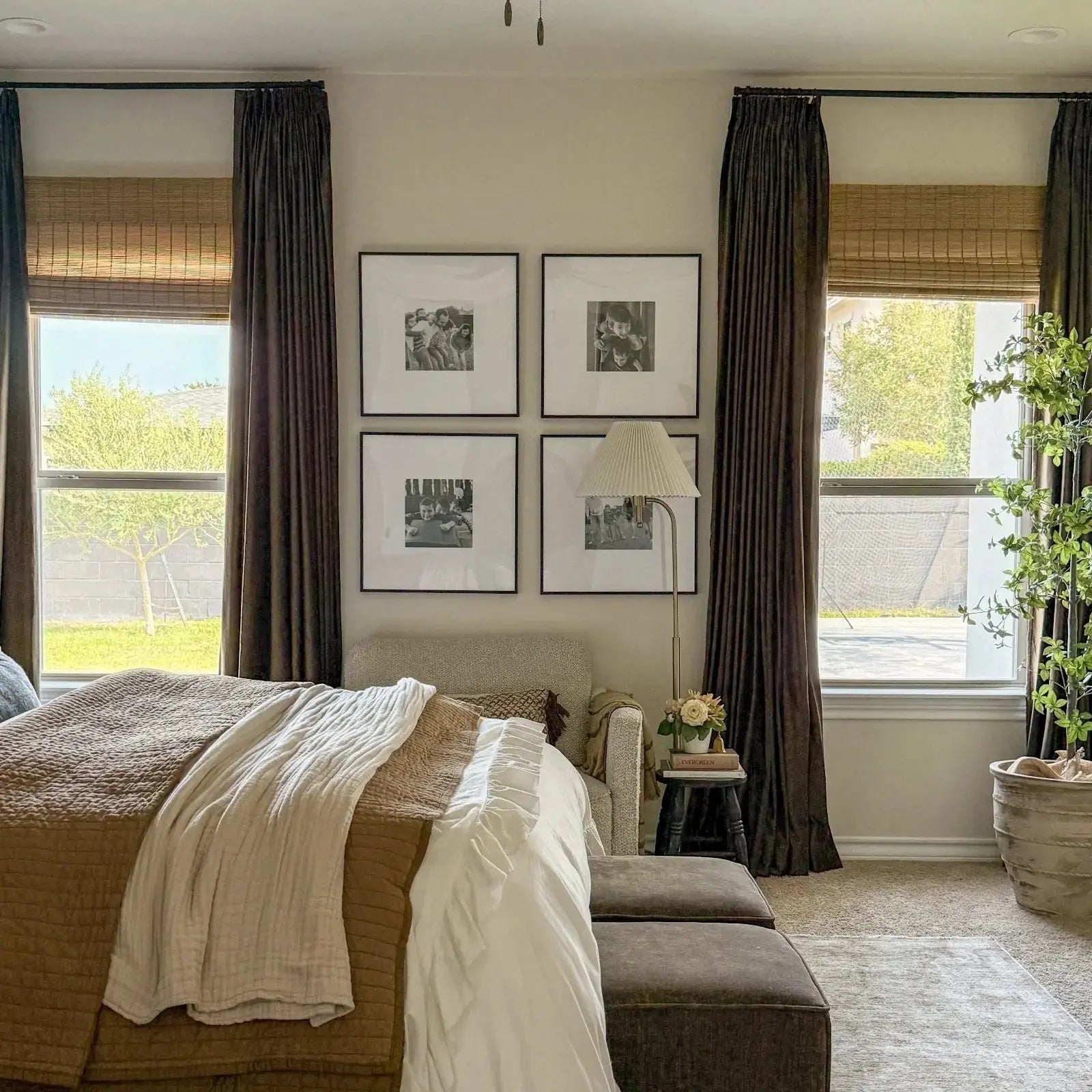 Modern bedroom with a brown quilted bed, layered white and beige throws, a gallery wall of four black and white family photos, large windows with dark brown velvet curtains and bamboo shades overlooking a green backyard, a pleated floor lamp, and a tall potted plant