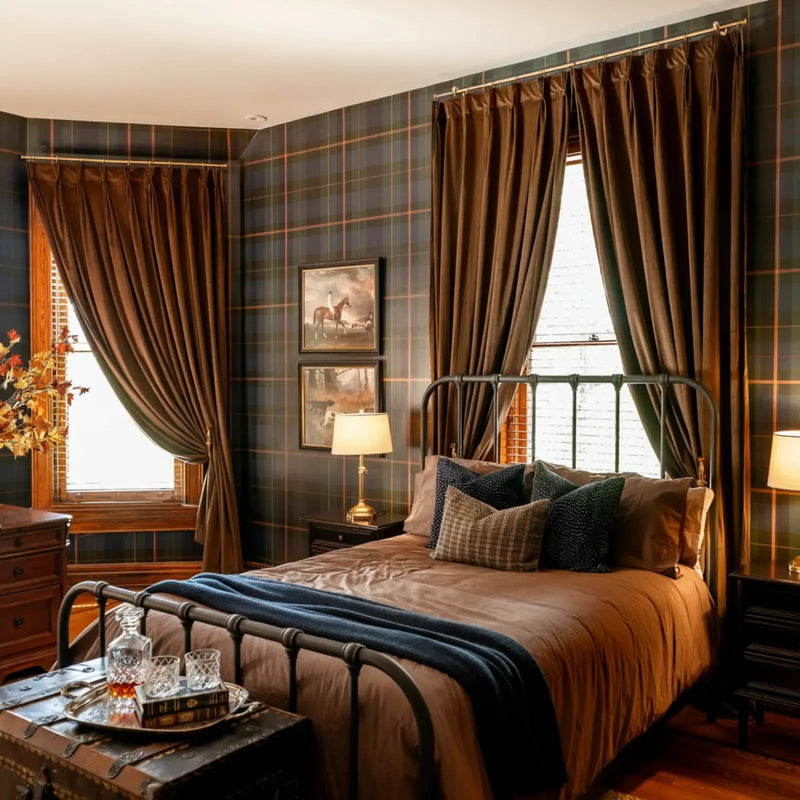 Photograph of a cozy, traditional bedroom featuring dark plaid wallpaper, rich brown velvet curtains, an iron bed with warm brown bedding and a contrasting blue throw, two equestrian art prints, and a vintage trunk with a decanter and crystal glasses