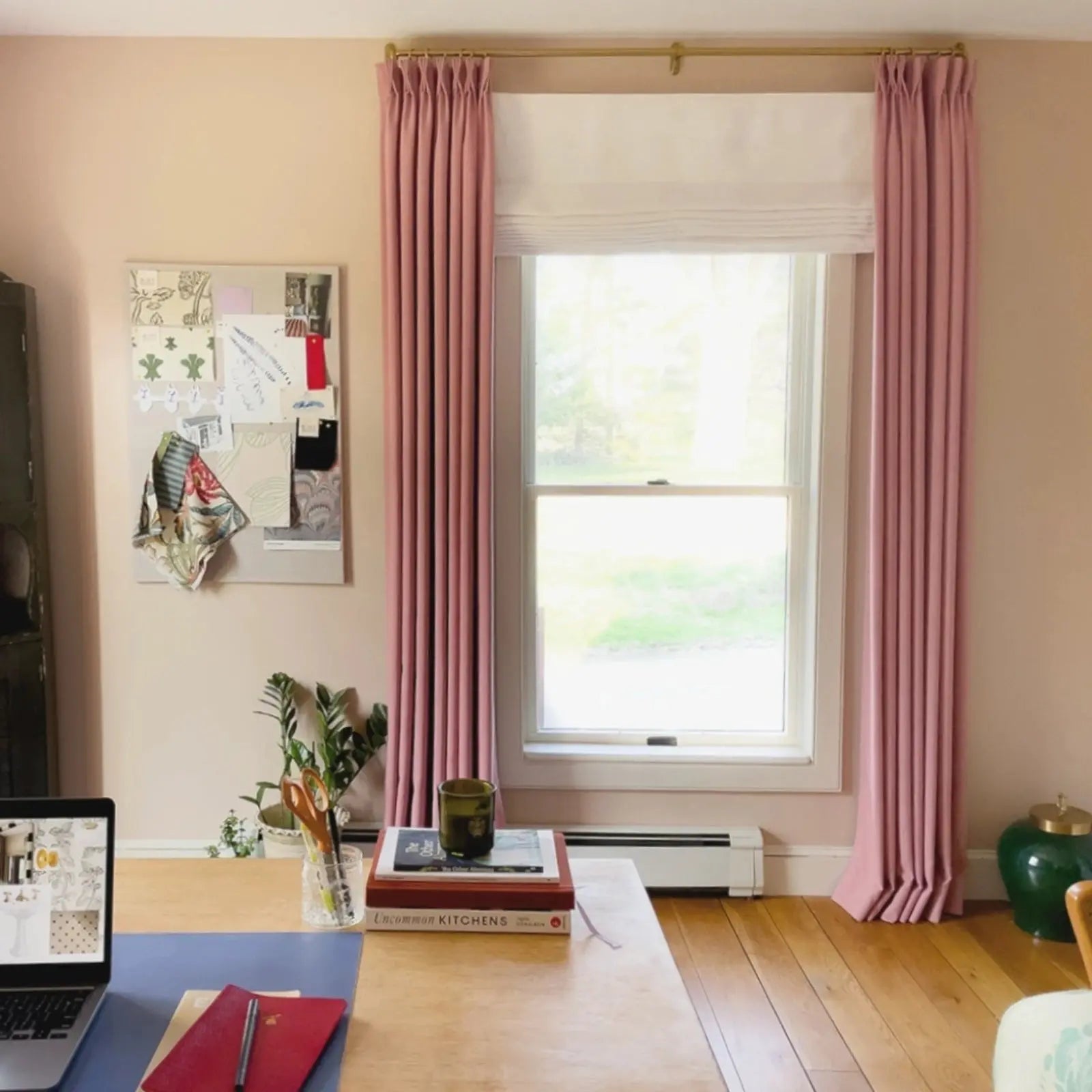Photography of a home office interior featuring a light wood desk with a laptop and red notebook in front of a window flanked by elegant pink pleated drapes on a gold rod, a design mood board with fabric swatches, and a vibrant green ceramic jar
