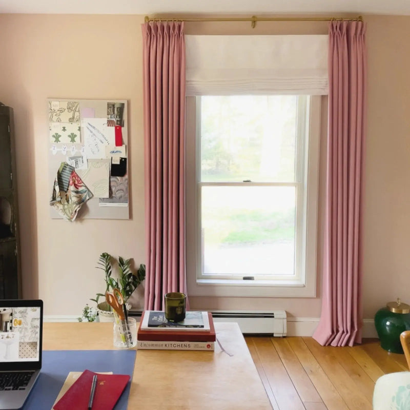 Photography of a home office interior featuring a light wood desk with a laptop and red notebook in front of a window flanked by elegant pink pleated drapes on a gold rod, a design mood board with fabric swatches, and a vibrant green ceramic jar