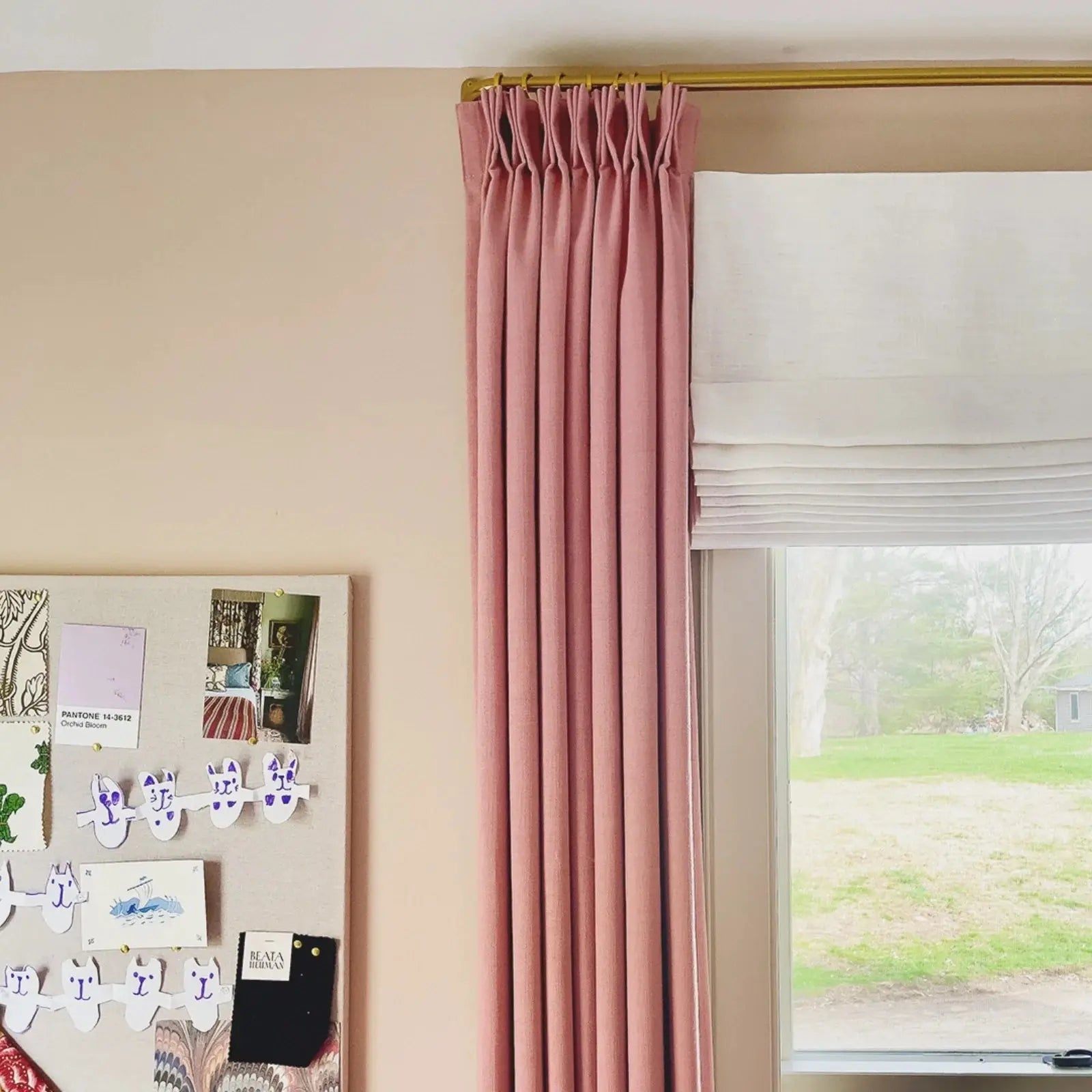 Interior view of a pink wall with a mood board displaying design swatches and paper cat chains beside a window featuring a pink pinch-pleat curtain on a gold rod and a white Roman blind overlooking a green backyard with a small grey shed