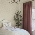 A serene child's bedroom showcasing a white bed with a pillow depicting a mouse on a swing and a quilt with red mushrooms, surrounded by soft patterned wallpaper featuring rabbits and an olive tree plant beside a blush pink velvet curtain