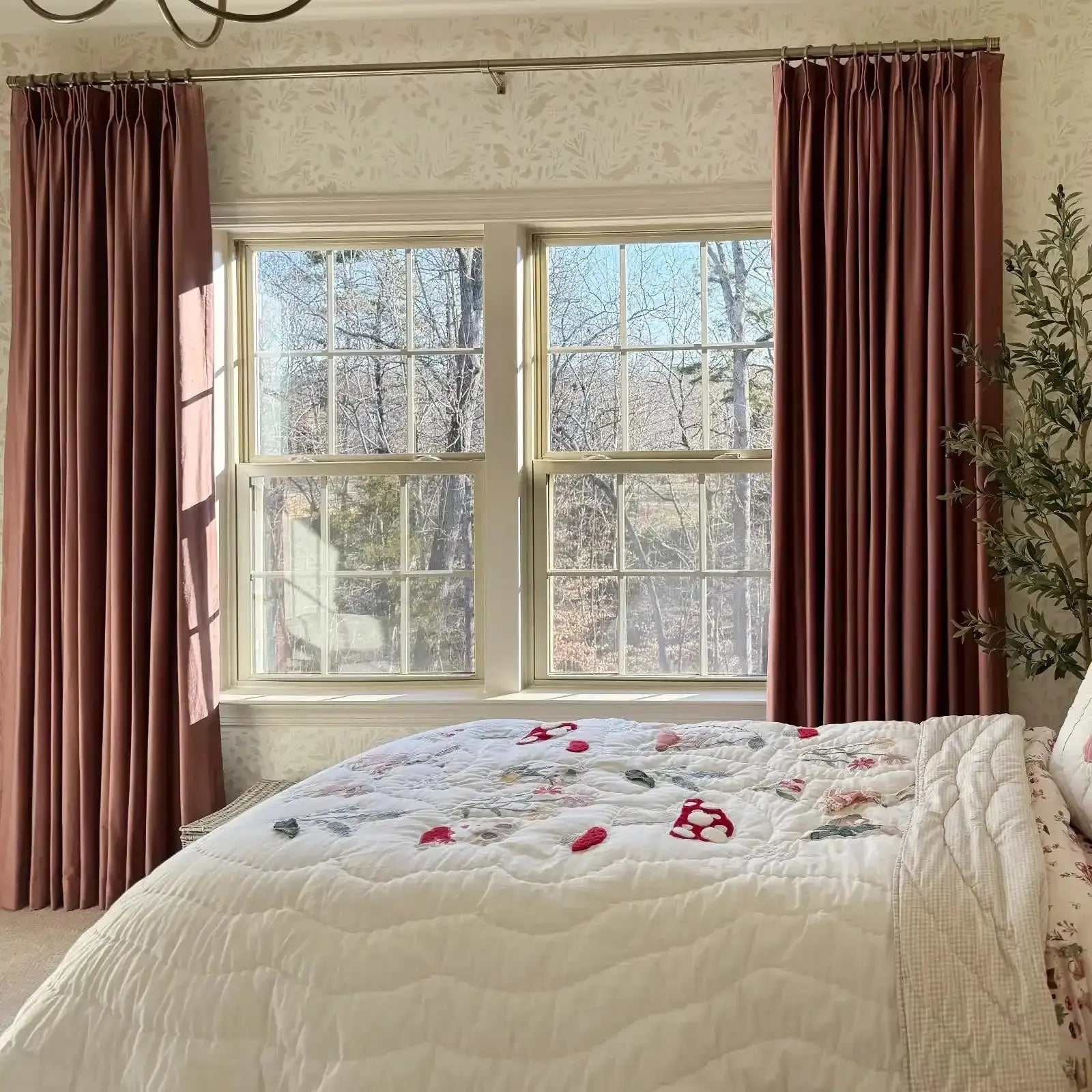 A cozy bedroom interior featuring a white quilted bedspread with red mushroom and floral appliques, dusty rose velvet curtains framing a window overlooking a bare winter forest, and light beige botanical wallpaper