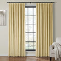 Photograph of a modern living room featuring two pleated beige curtains on a black rod framing a window with black grid panes, a minimalist gray tree artwork on the wall, and a light gray armchair with a white fringed throw