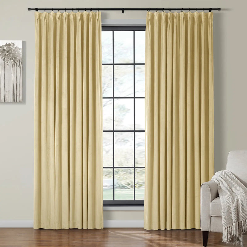 Photograph of a modern living room featuring two pleated beige curtains on a black rod framing a window with black grid panes, a minimalist gray tree artwork on the wall, and a light gray armchair with a white fringed throw