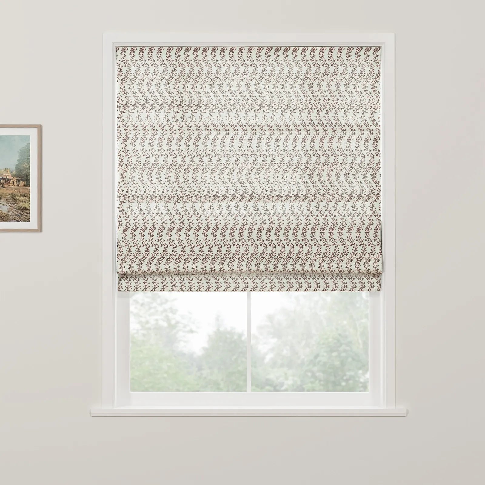 A white-framed window featuring a cream roman blind with a repetitive brown vine and leaf pattern, revealing a blurred green outdoor tree view, next to a small framed landscape painting on a white wall