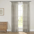 Photograph of a bright room featuring a window dressed with pleated cream curtains displaying a subtle green-brown leafy pattern on a black rod, alongside a light wood mid-century modern dresser with brass knobs and a framed botanical art print depicting yellow flowers