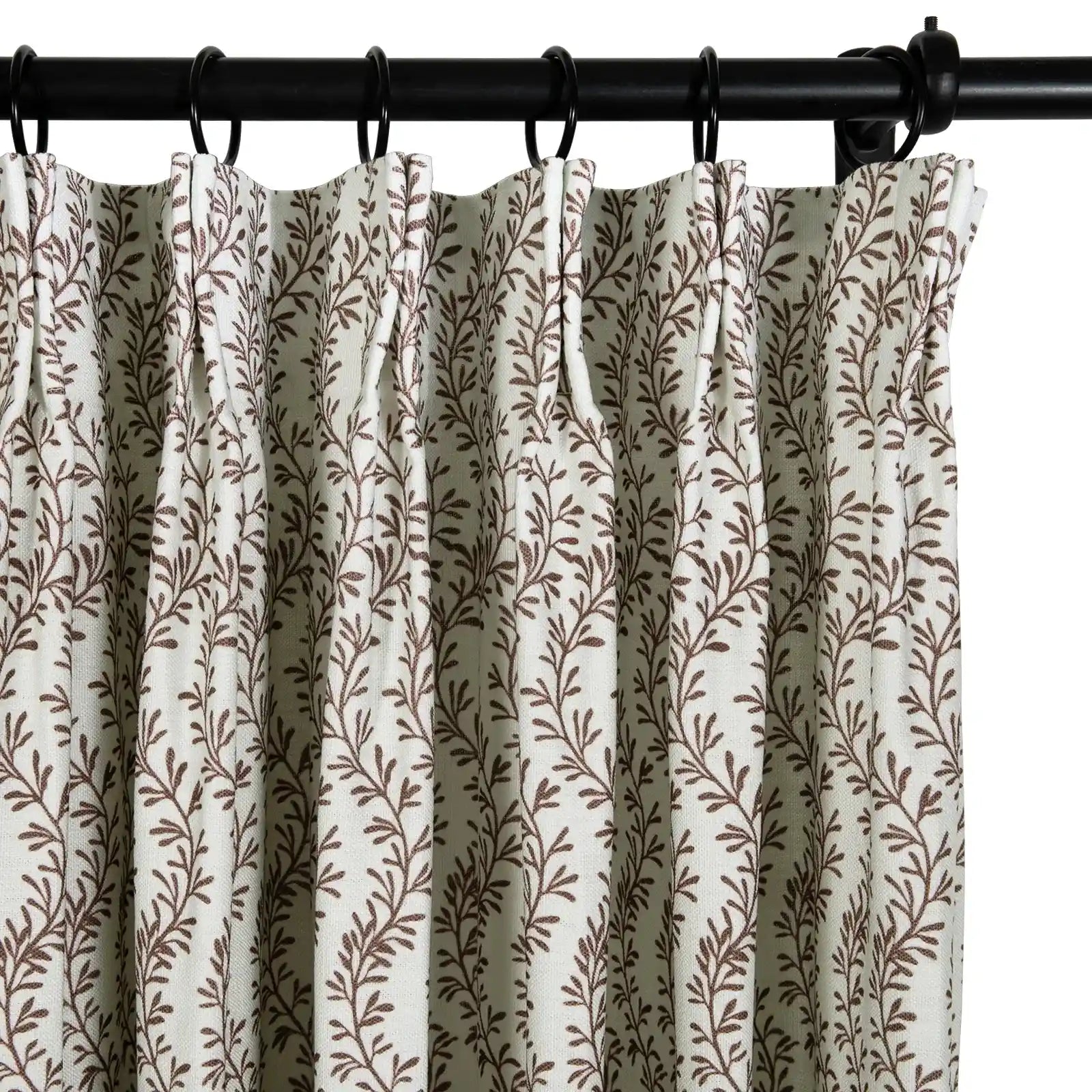 Textile image of an off-white pleated curtain adorned with a dense brown leafy vine pattern, suspended by black rings on a dark metal rod
