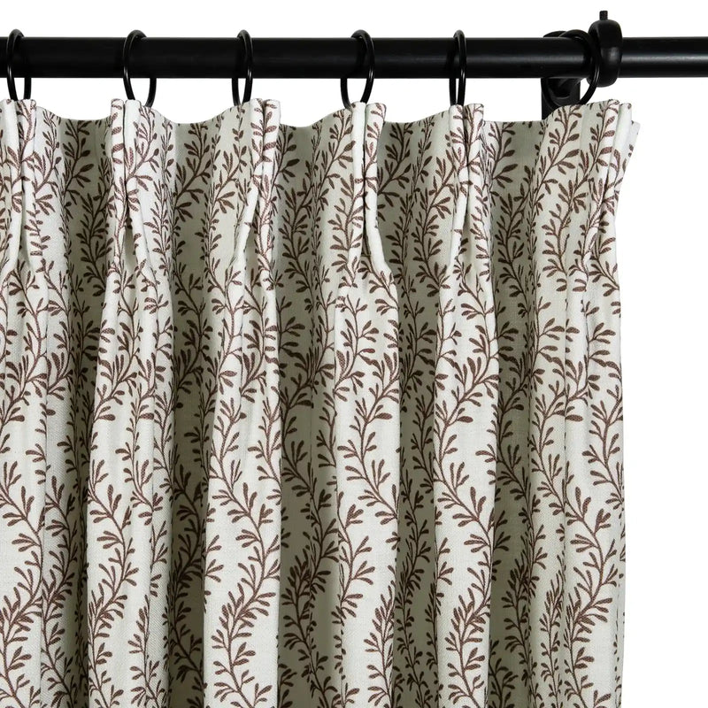 Textile image of an off-white pleated curtain adorned with a dense brown leafy vine pattern, suspended by black rings on a dark metal rod